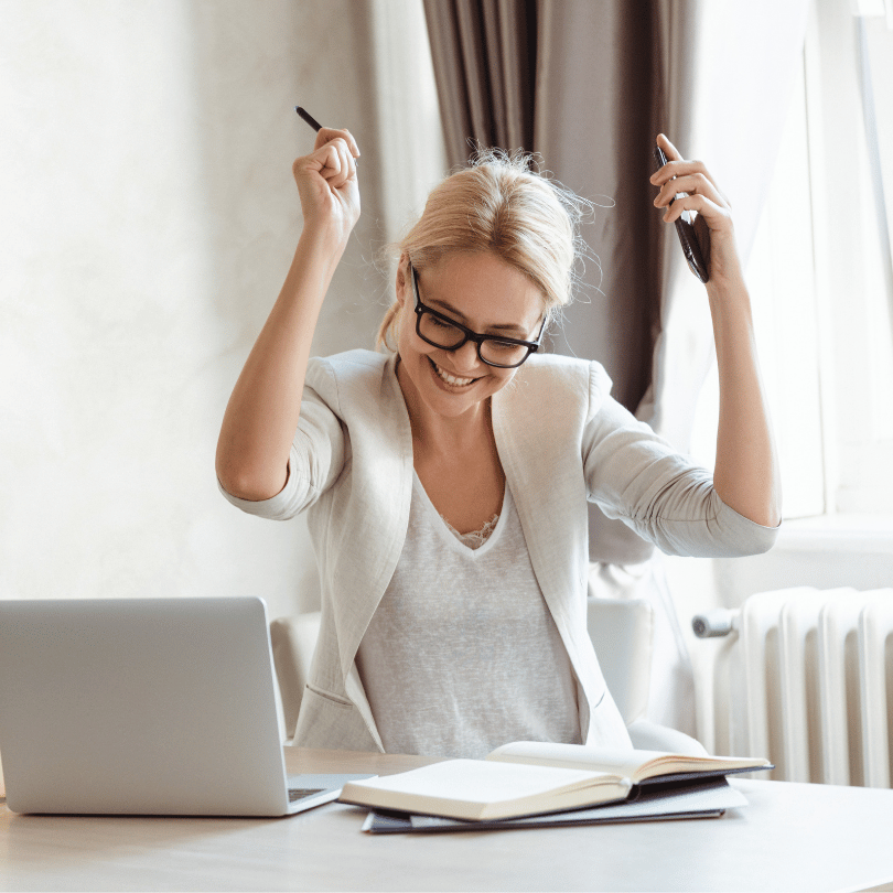 Woman sitting at desk and raising arms in air with success. She is holding a phone in one hand and a pen in the other. She is smiling.